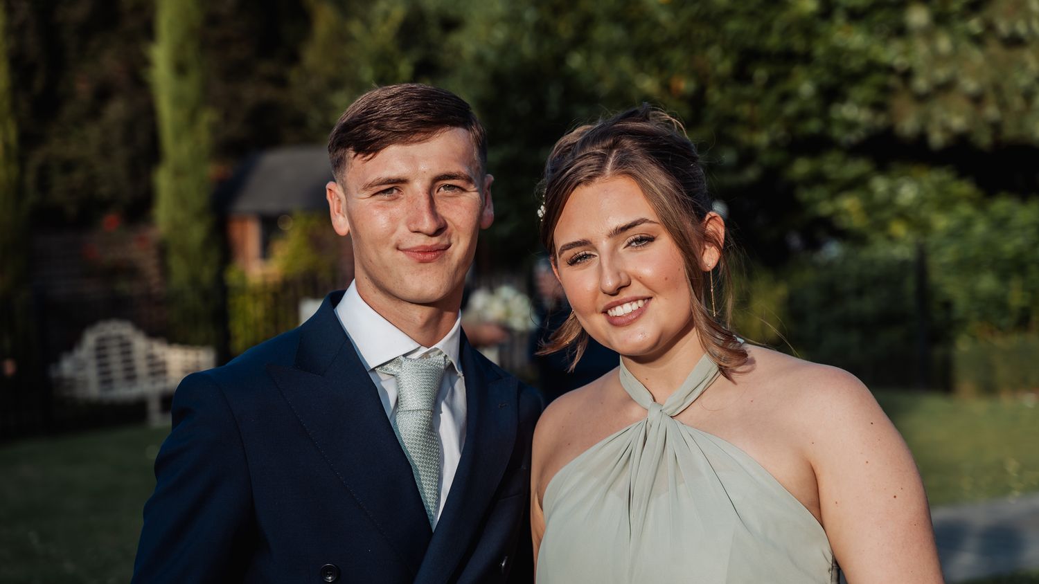Smiling couple dressed formally in a garden setting, man in navy suit and woman in sage green halter dress.