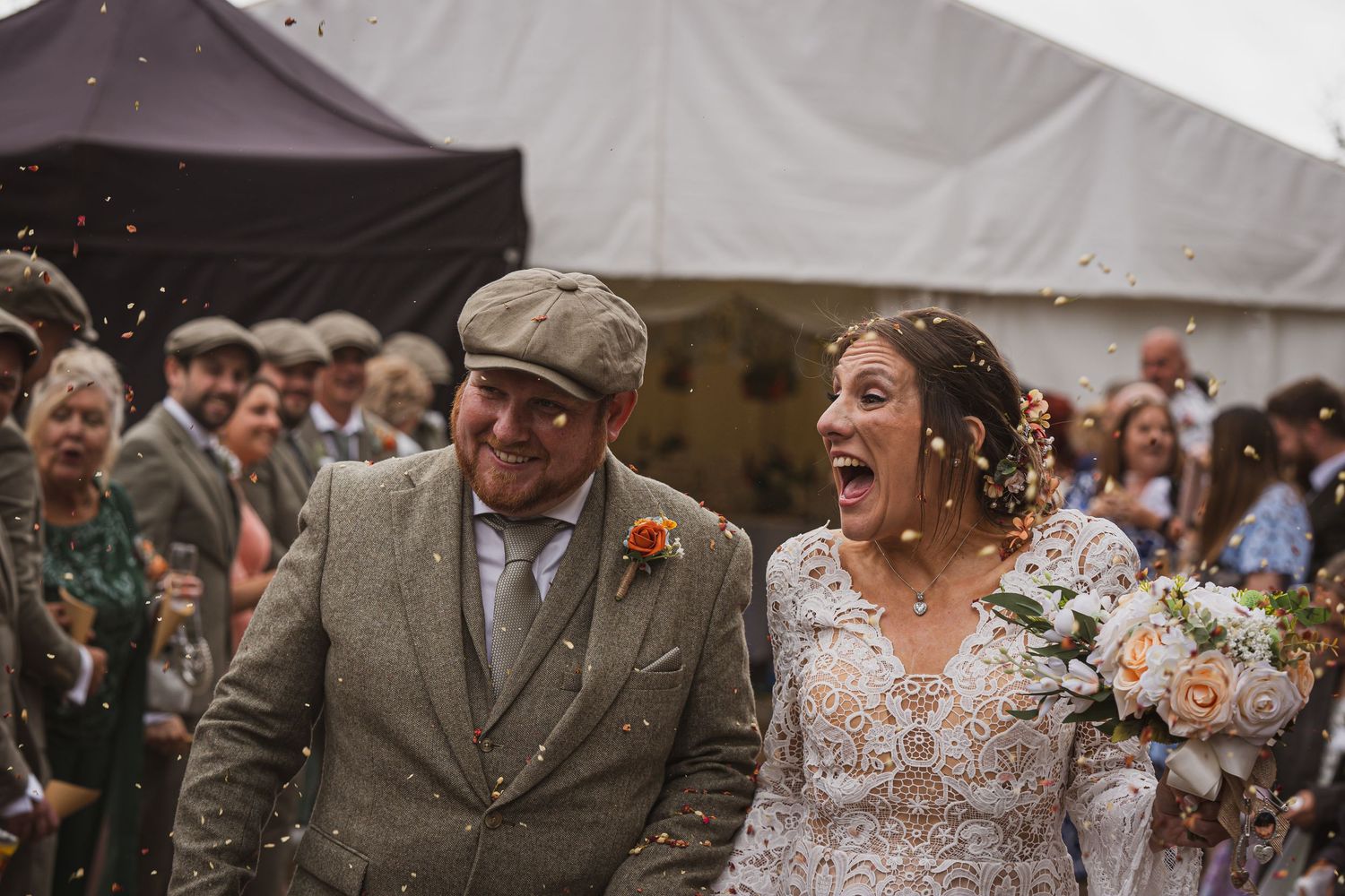 Joyful bride and groom laughing in confetti shower at outdoor wedding celebration with guests.