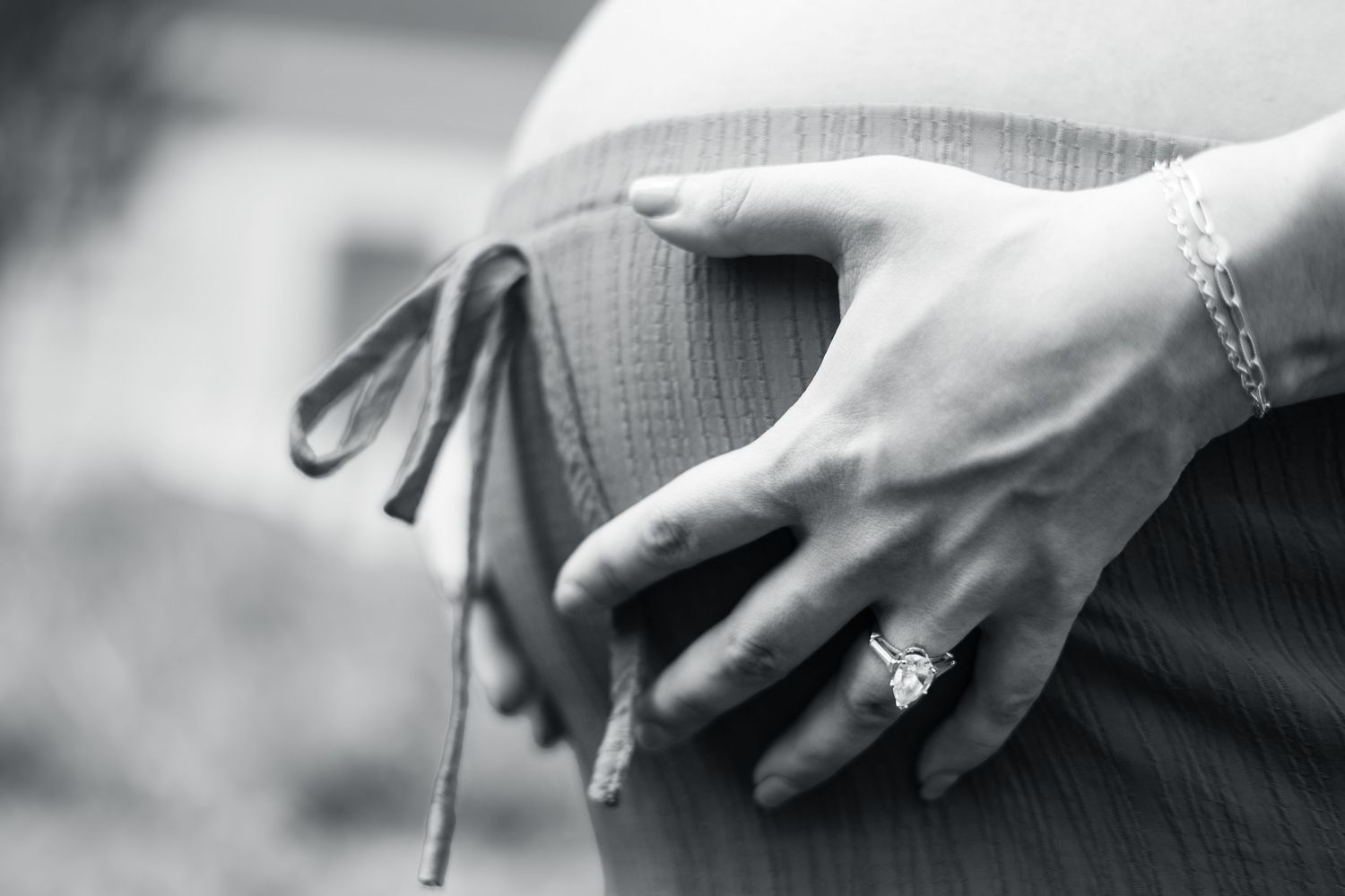 Black and white close-up of a pregnant woman cradling her baby bump, wearing a diamond ring and bracelet.