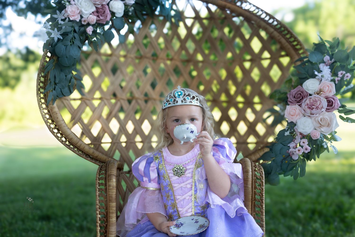 A little girl in a purple Rapunzel princess dress and tiara sits in a wicker throne chair with floral decorations outdoors.