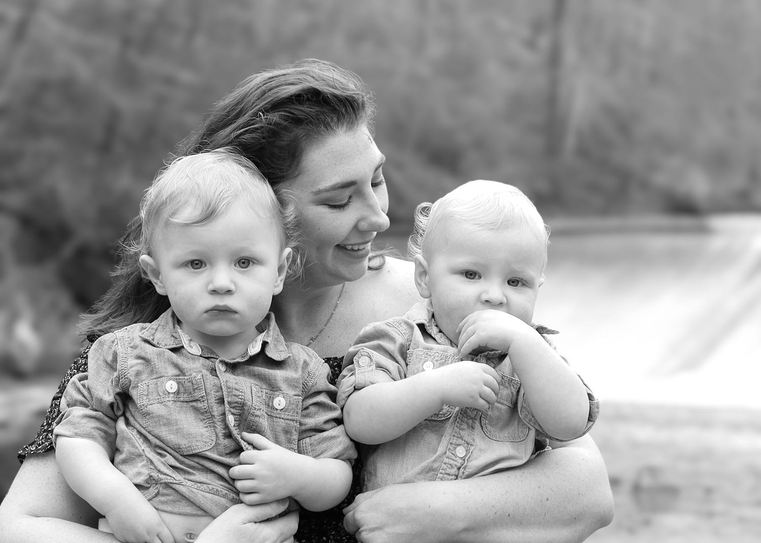 Black and white photo of a smiling mother holding two young toddlers outdoors near a waterfall.