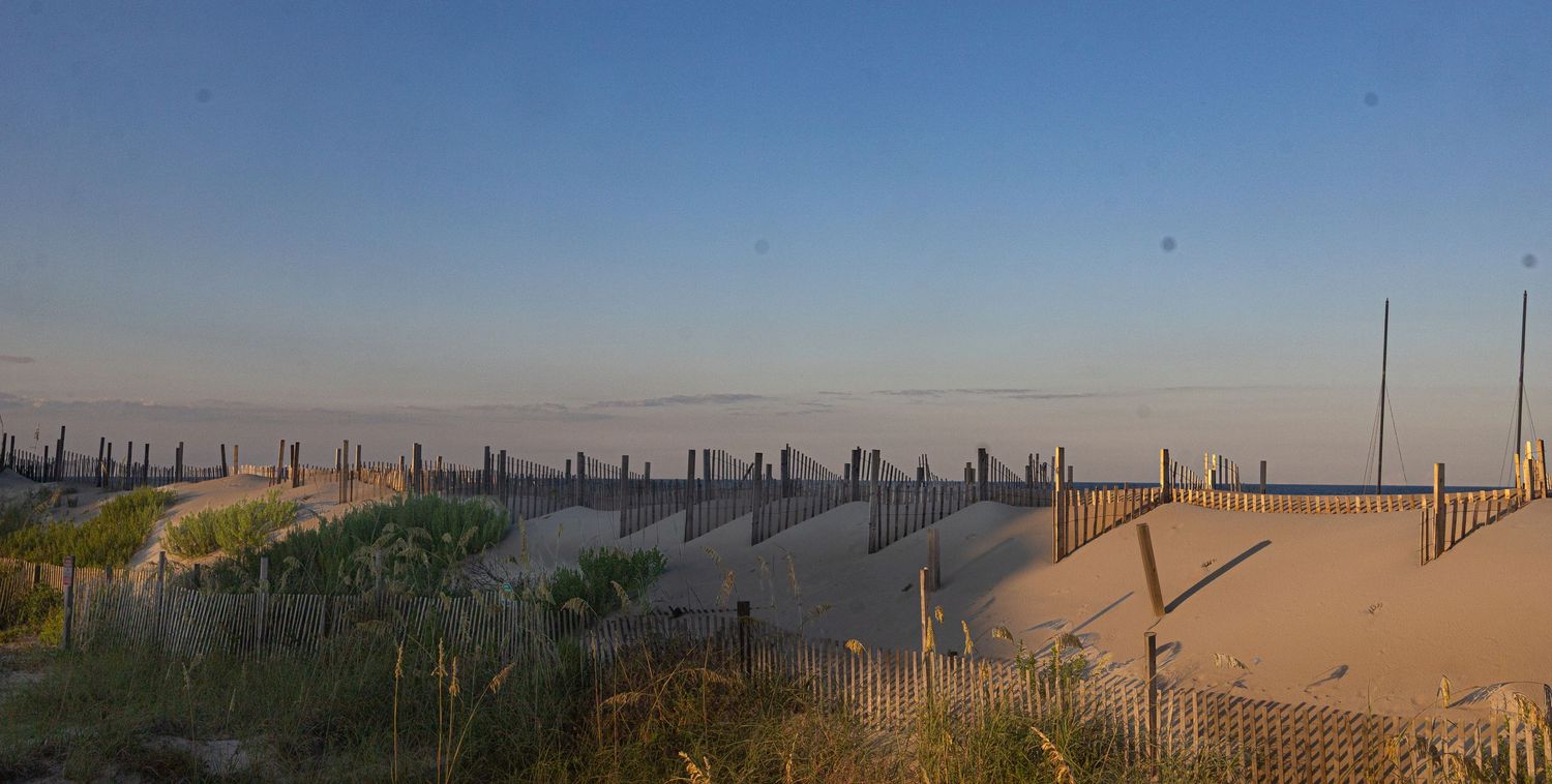 Sandy beach dunes with wooden snow fencing at sunset, calm ocean horizon under a clear blue sky.