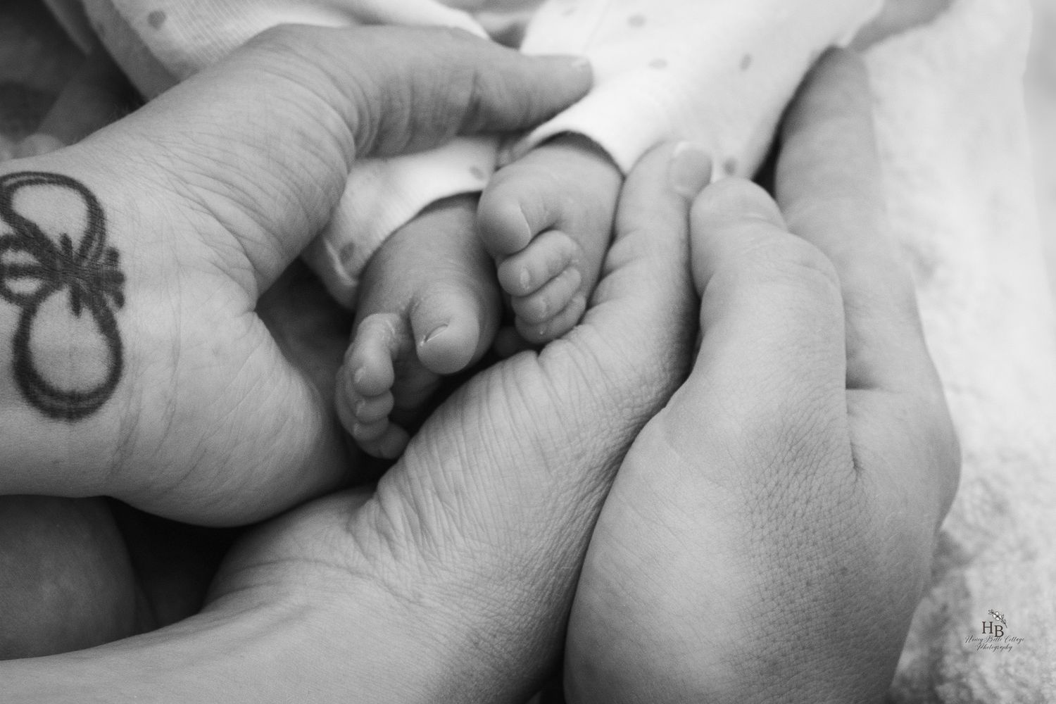 Black and white photo of adult hands cradling tiny newborn baby feet, with an infinity tattoo visible.