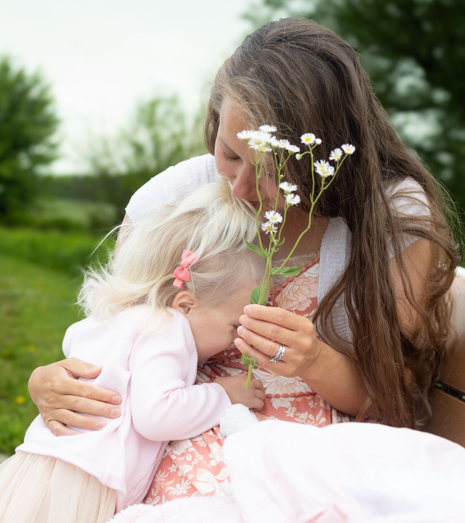 A mother with long brown hair hugs a young blonde girl while holding white wildflowers outdoors.
