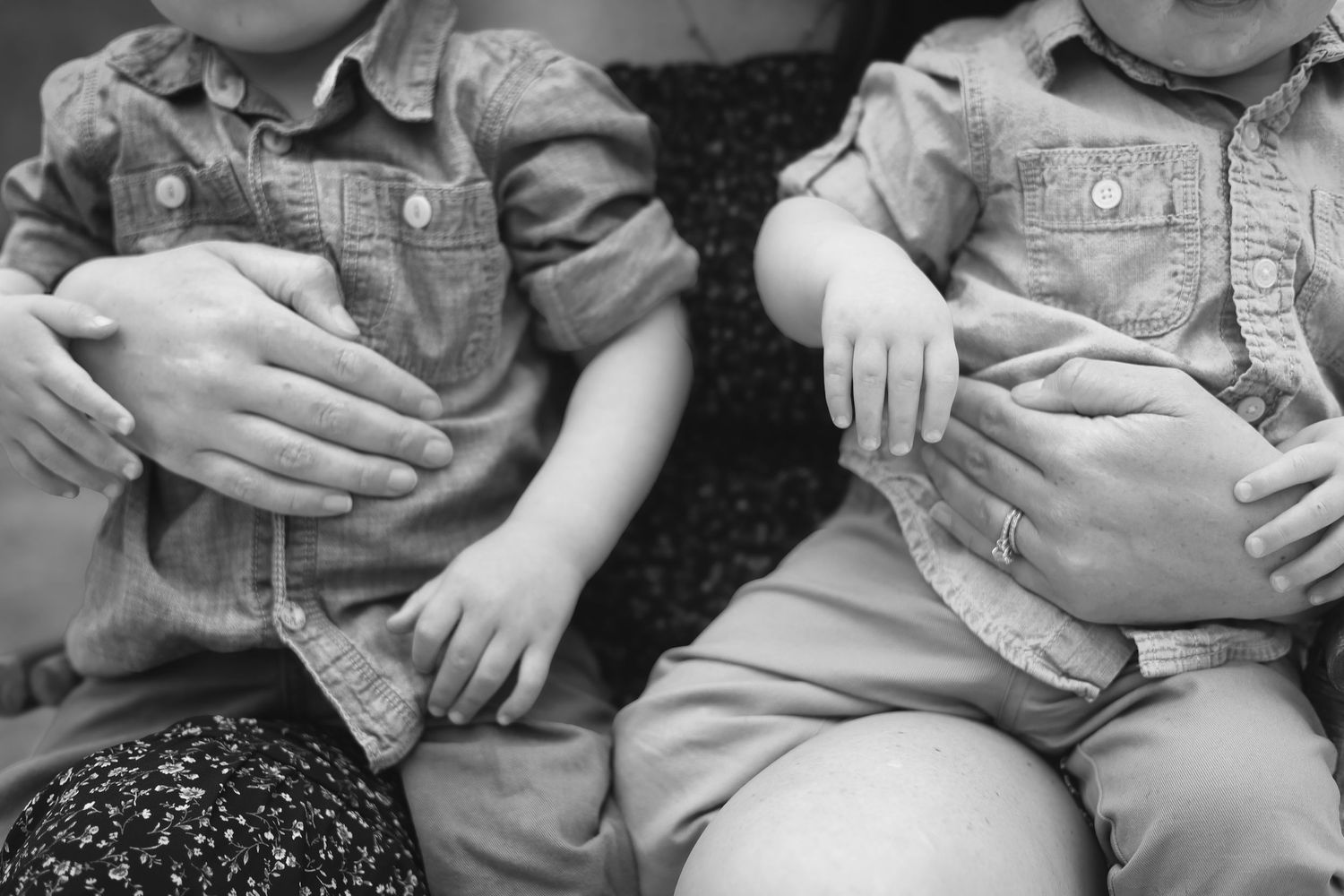 Black and white photo of twin toddlers in matching denim shirts sitting side by side with adult hands resting gently.