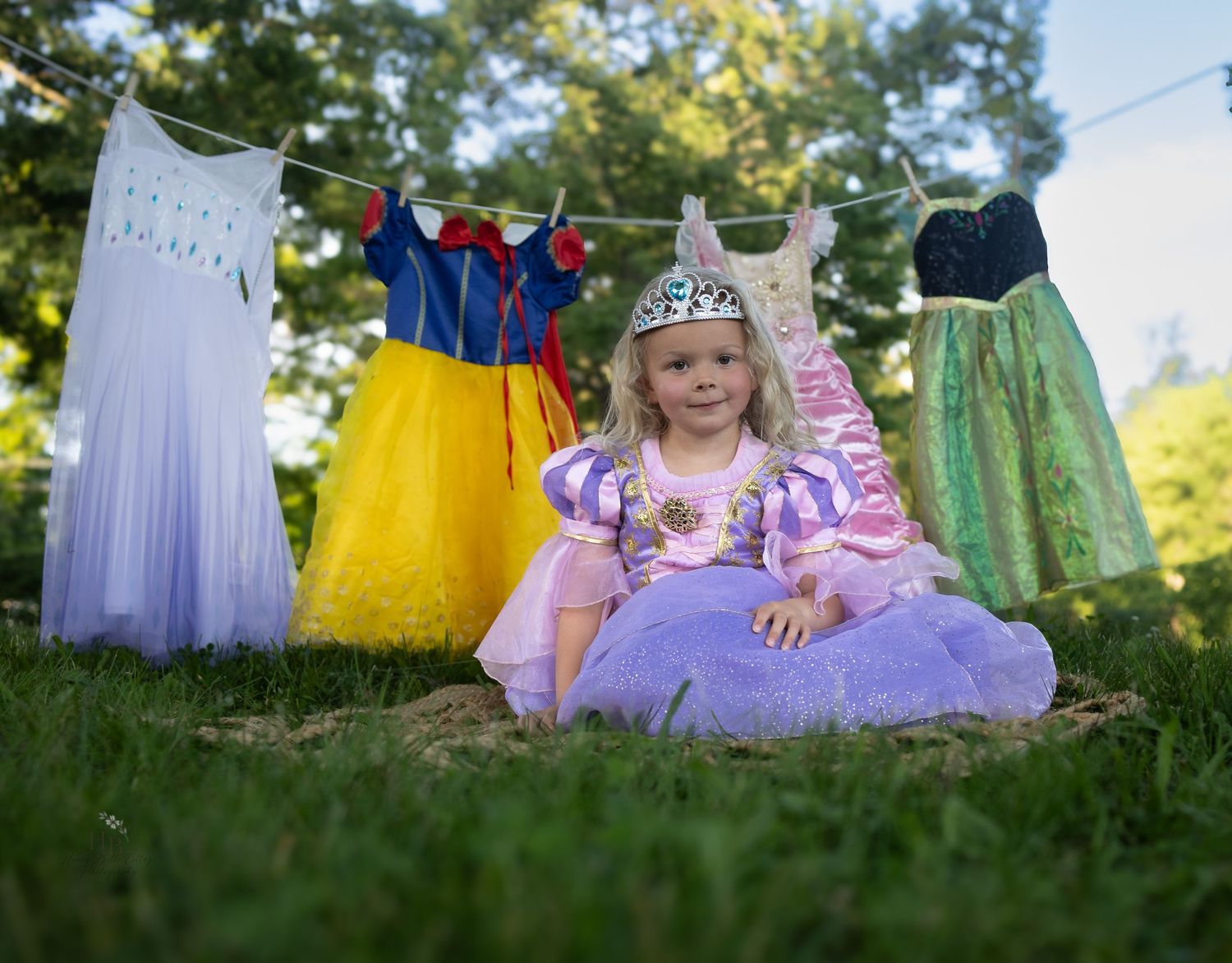 A young girl in a purple Rapunzel princess dress sits on grass with Disney princess costumes hanging on a clothesline behind her.