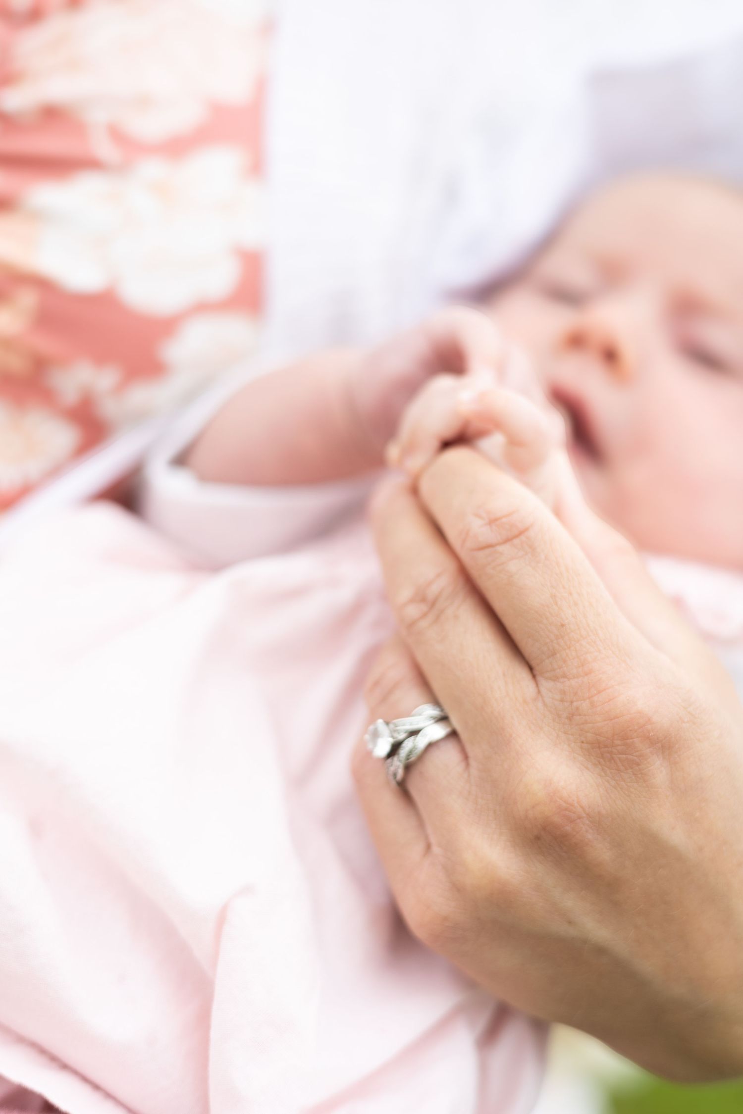 A mother's hand with diamond rings gently holds a sleeping newborn baby's tiny fist in pink clothing.