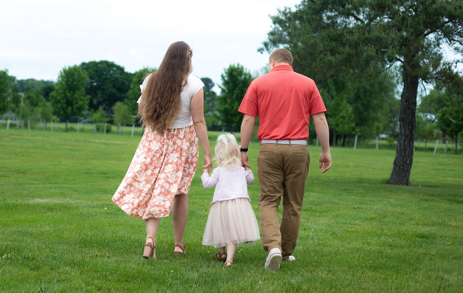 Family of three walking hand in hand across lush green park, viewed from behind.