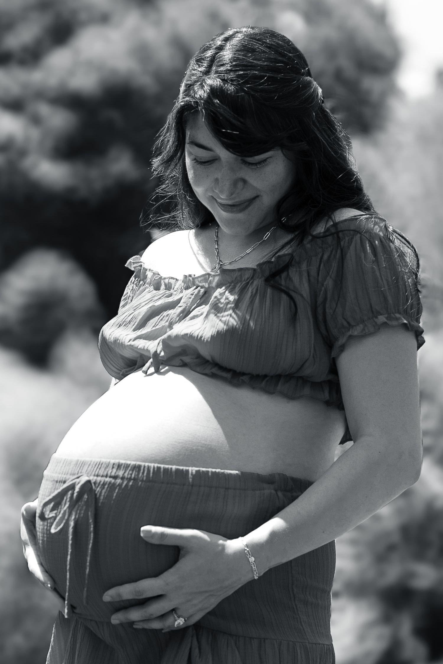 Black and white photo of a smiling pregnant woman cradling her baby bump outdoors.