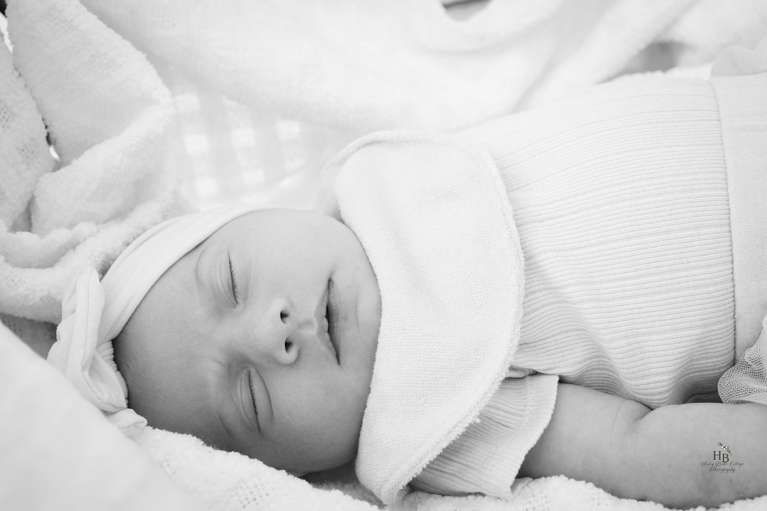 Black and white photo of a peaceful sleeping newborn baby wrapped in soft white blankets wearing a white hat.