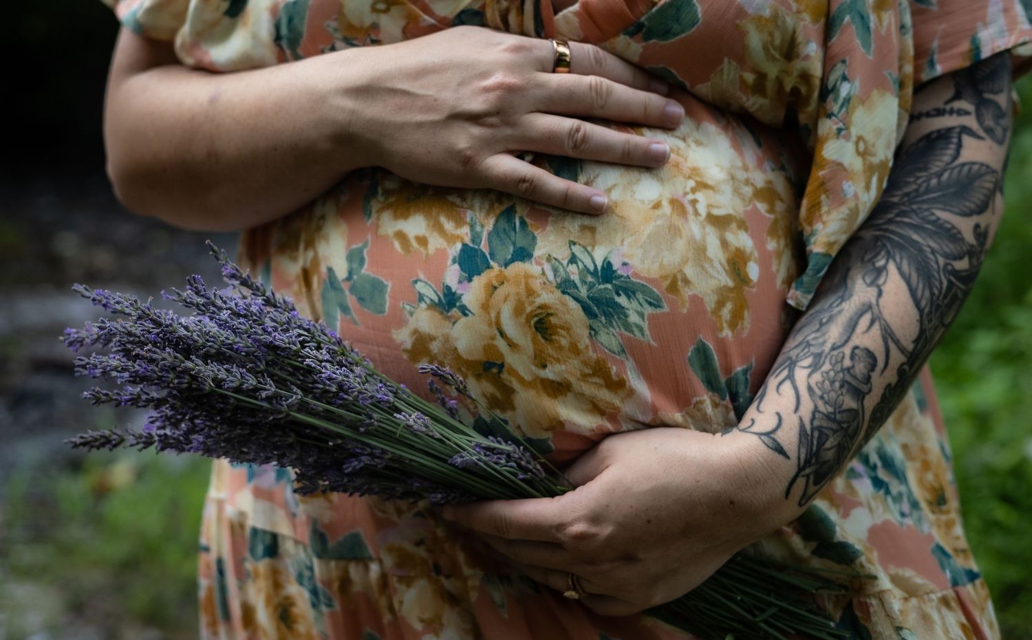 Pregnant woman in floral dress holding lavender bouquet, showcasing tattoo sleeve, maternity photography outdoors.