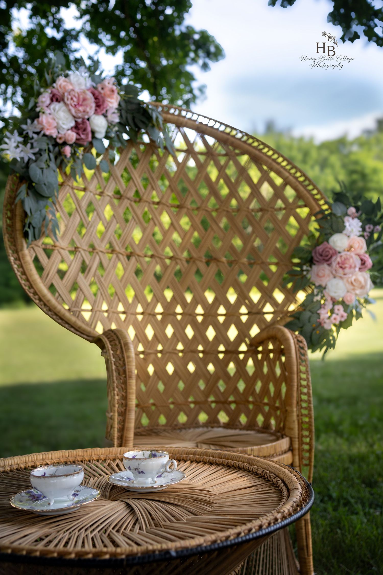 Elegant wicker peacock chair adorned with pink roses, two porcelain teacups on rattan table, set in lush green garden.