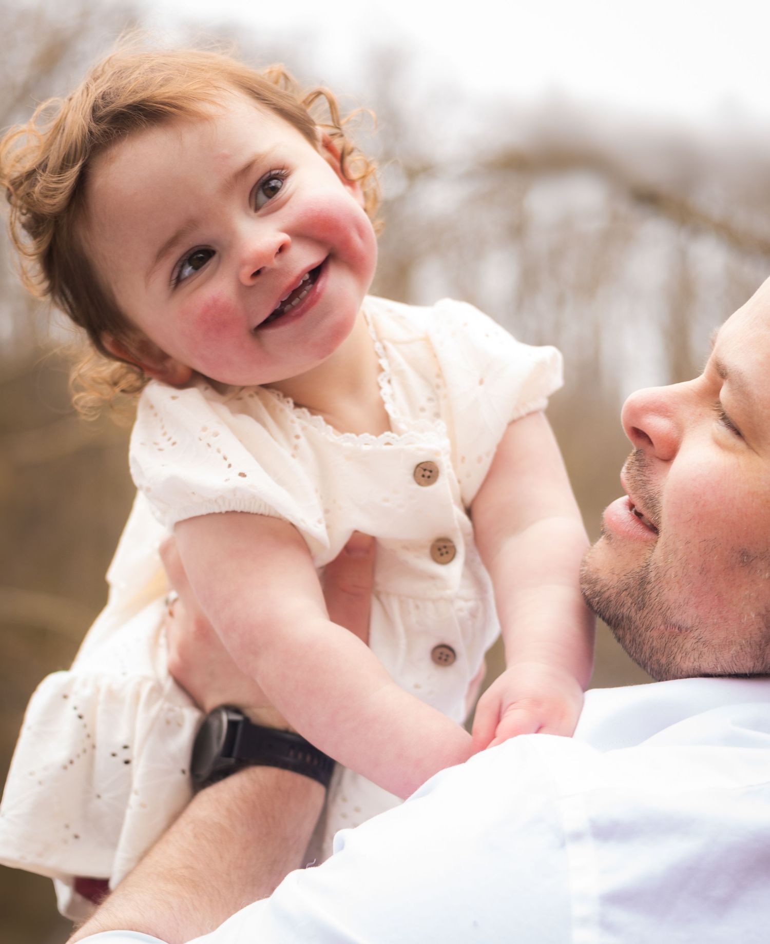 A joyful toddler girl in a white dress being lifted by her smiling father outdoors in a bright spring setting.