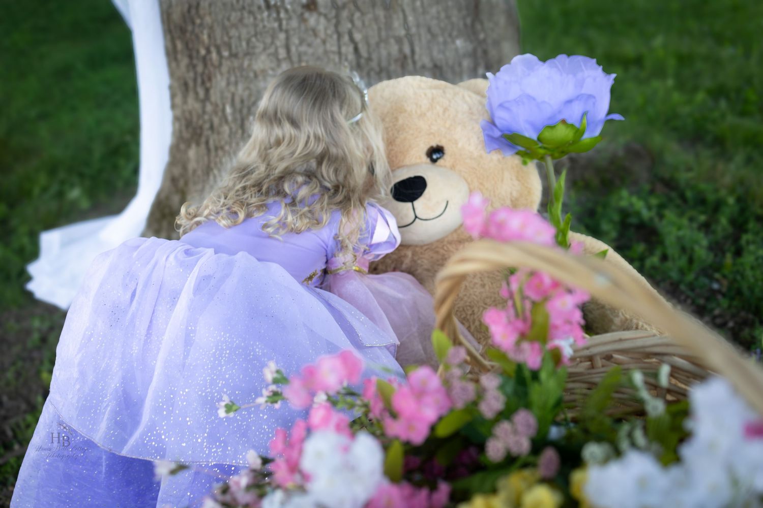 Young girl in purple dress hugging large teddy bear near tree surrounded by colorful flowers outdoors.