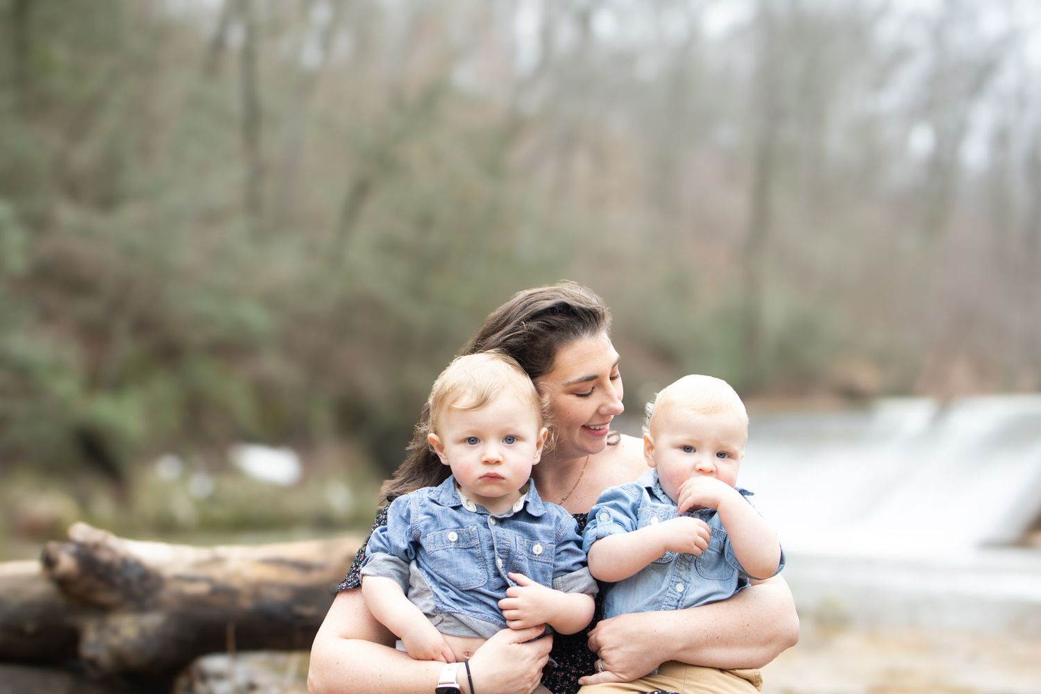 A mother holds two young toddlers in matching blue shirts near a serene waterfall in a wooded setting.