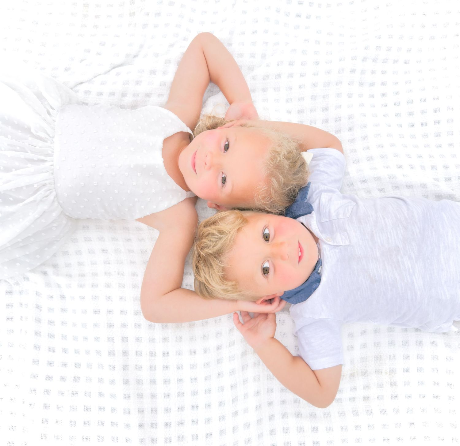 Two young blonde children lying head-to-head on a white surface, wearing white outfits, viewed from above.