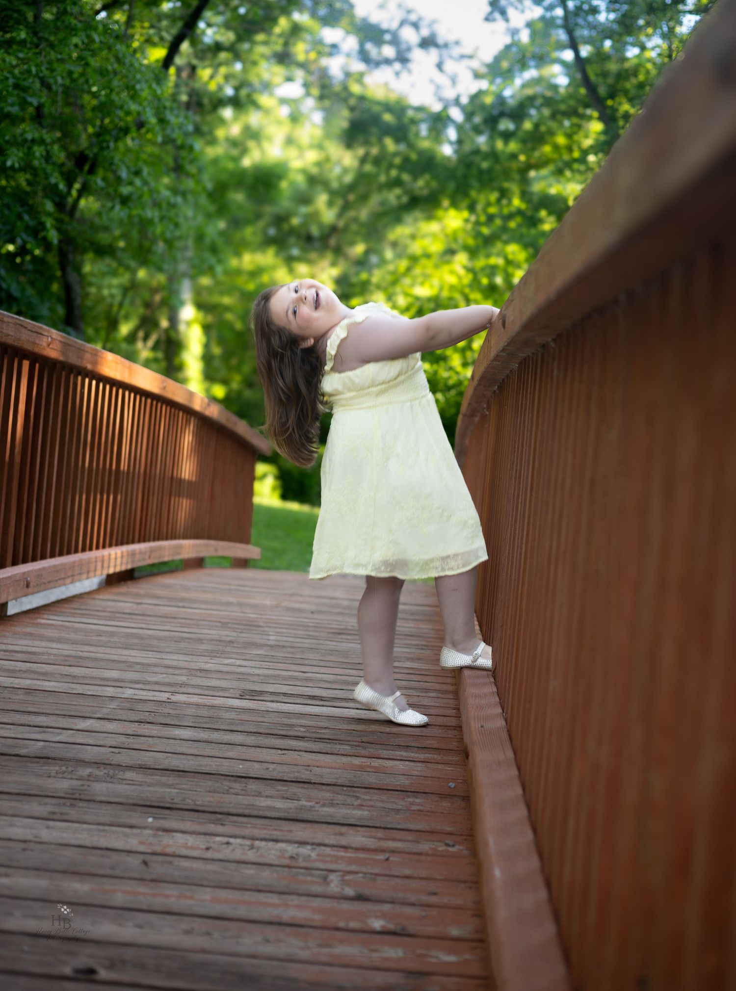 A joyful young girl in a yellow dress twirls on a wooden bridge surrounded by lush green trees.