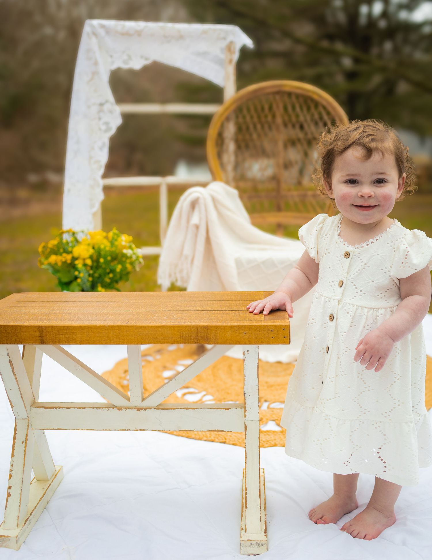 Smiling toddler girl in white dress standing barefoot beside a wooden table outdoors with yellow flowers.