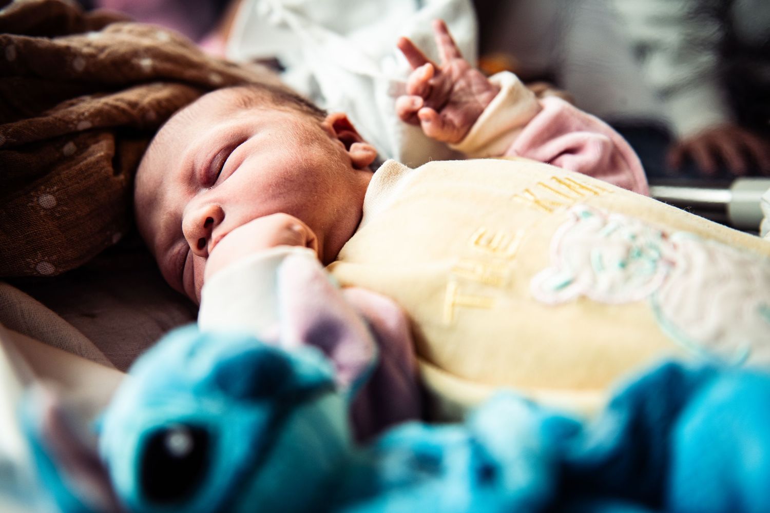 Newborn baby sleeping peacefully in yellow onesie next to a blue stuffed animal toy.