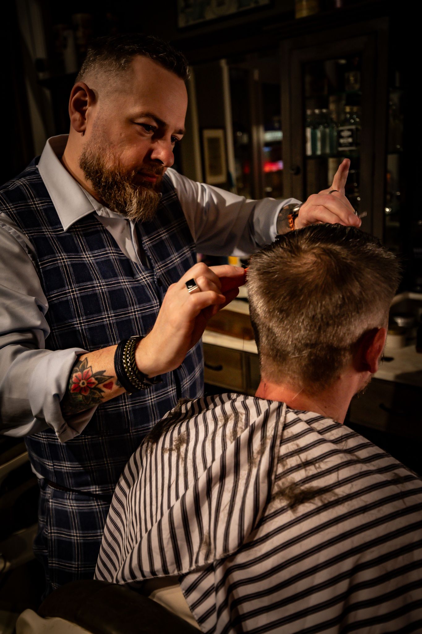 Tattooed barber in plaid vest cuts client's hair with scissors in a dark, moody barbershop setting.