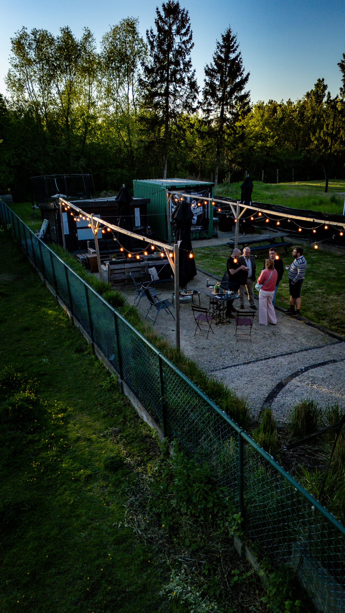 Outdoor evening gathering with string lights, guests socializing on a gravel patio surrounded by lush green trees and fencing.