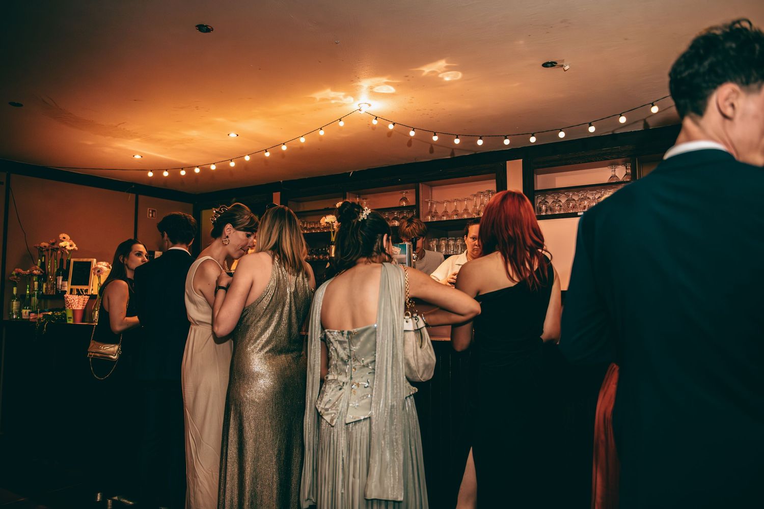 Wedding guests in elegant gowns socializing at a warmly lit bar during a reception.