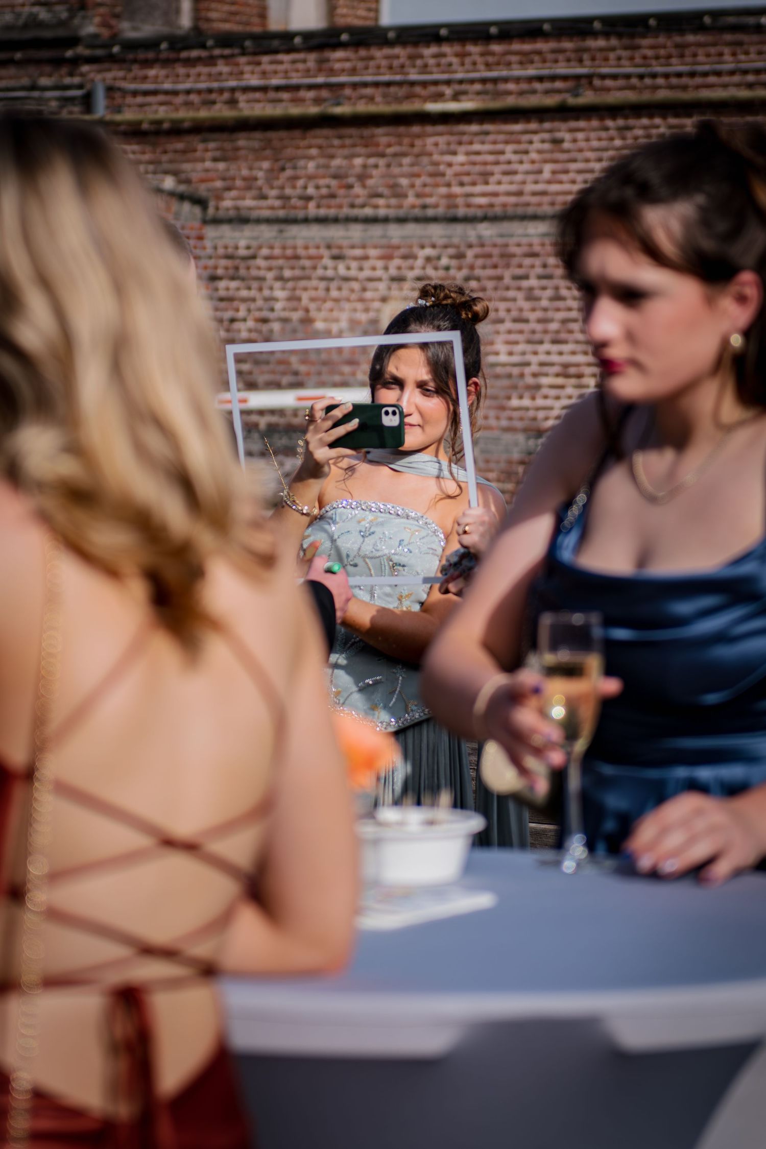Young women in formal dresses at an outdoor event, one taking a selfie in a mirror while another holds champagne.