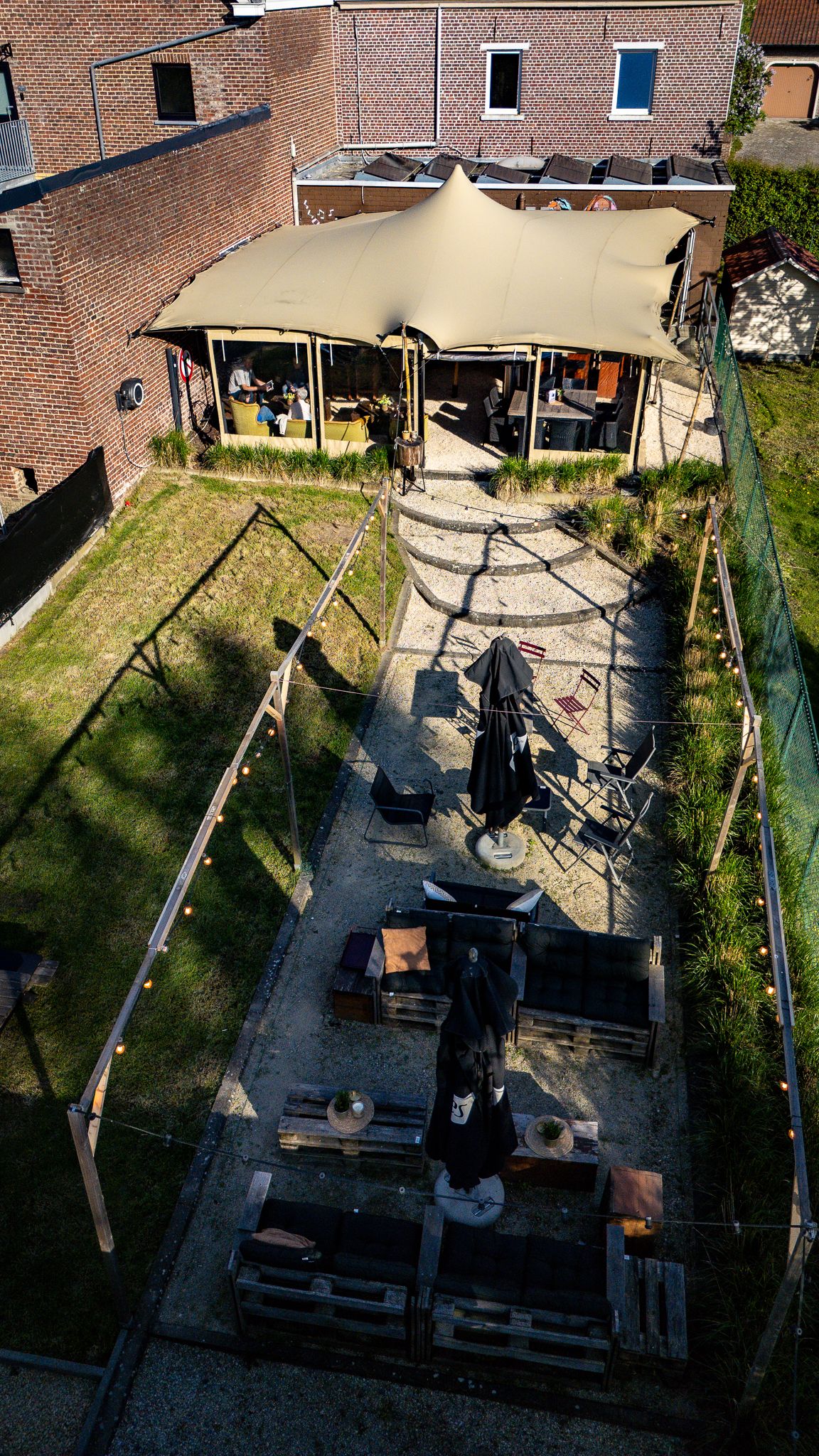 Aerial view of a backyard patio with a large tan canopy, string lights, pallet furniture, and a black umbrella.