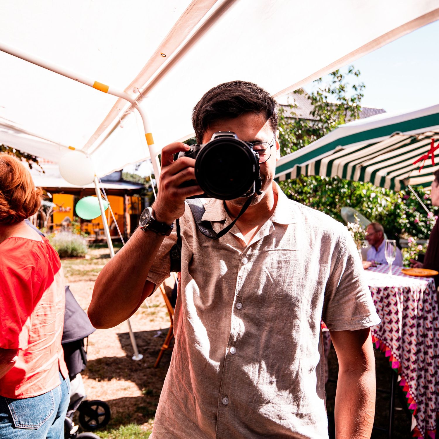 Man photographing with DSLR camera at outdoor market under striped tent on sunny day.