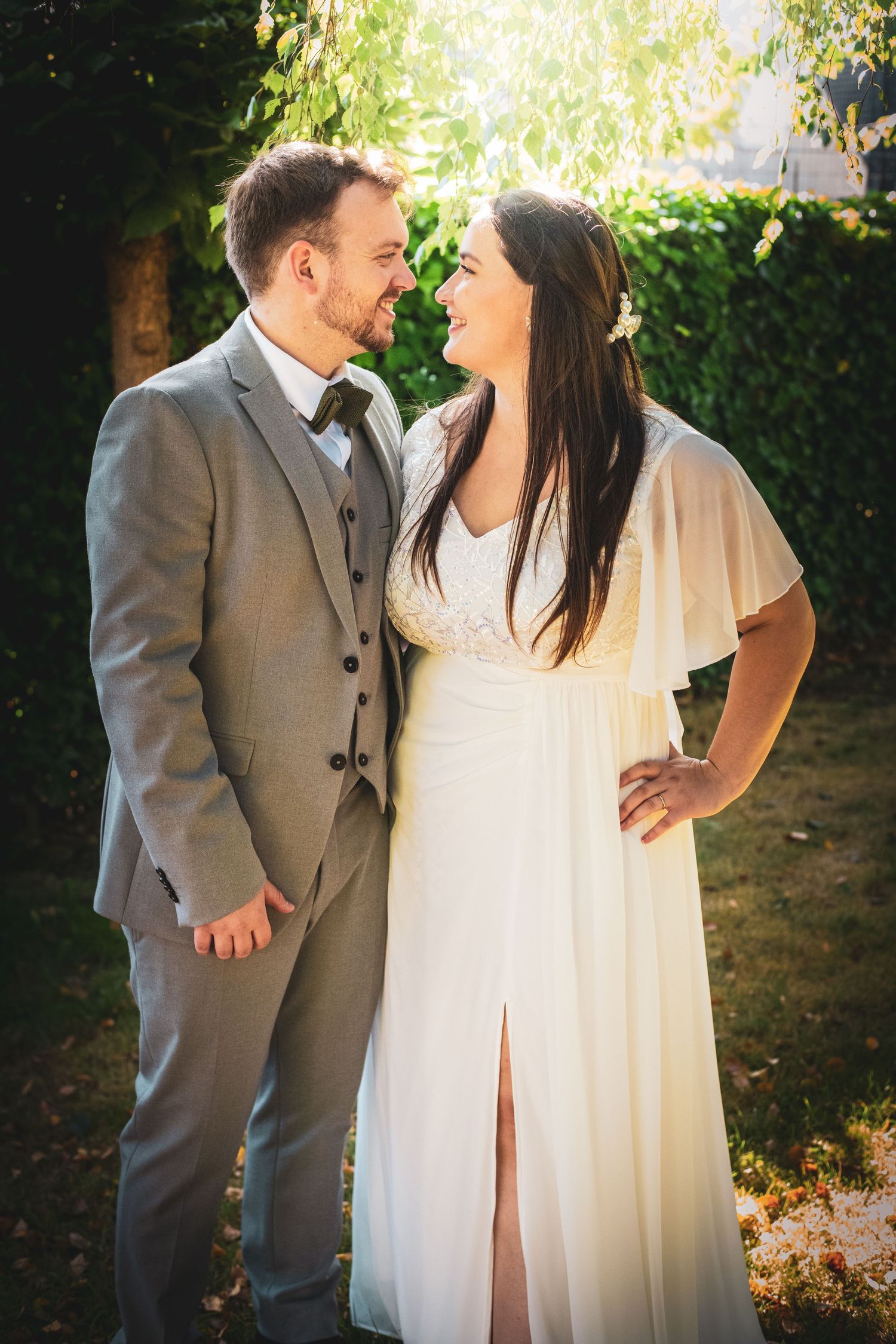A smiling bride in a white dress and groom in a gray suit share a romantic moment outdoors on their wedding day.
