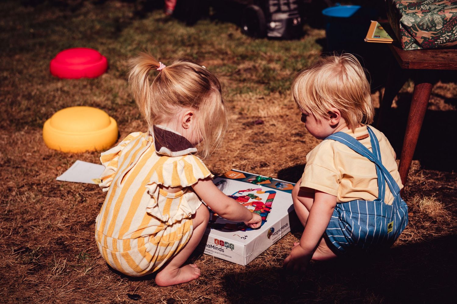 Two young toddlers in yellow outfits sit on grass playing with a colorful board game outdoors.