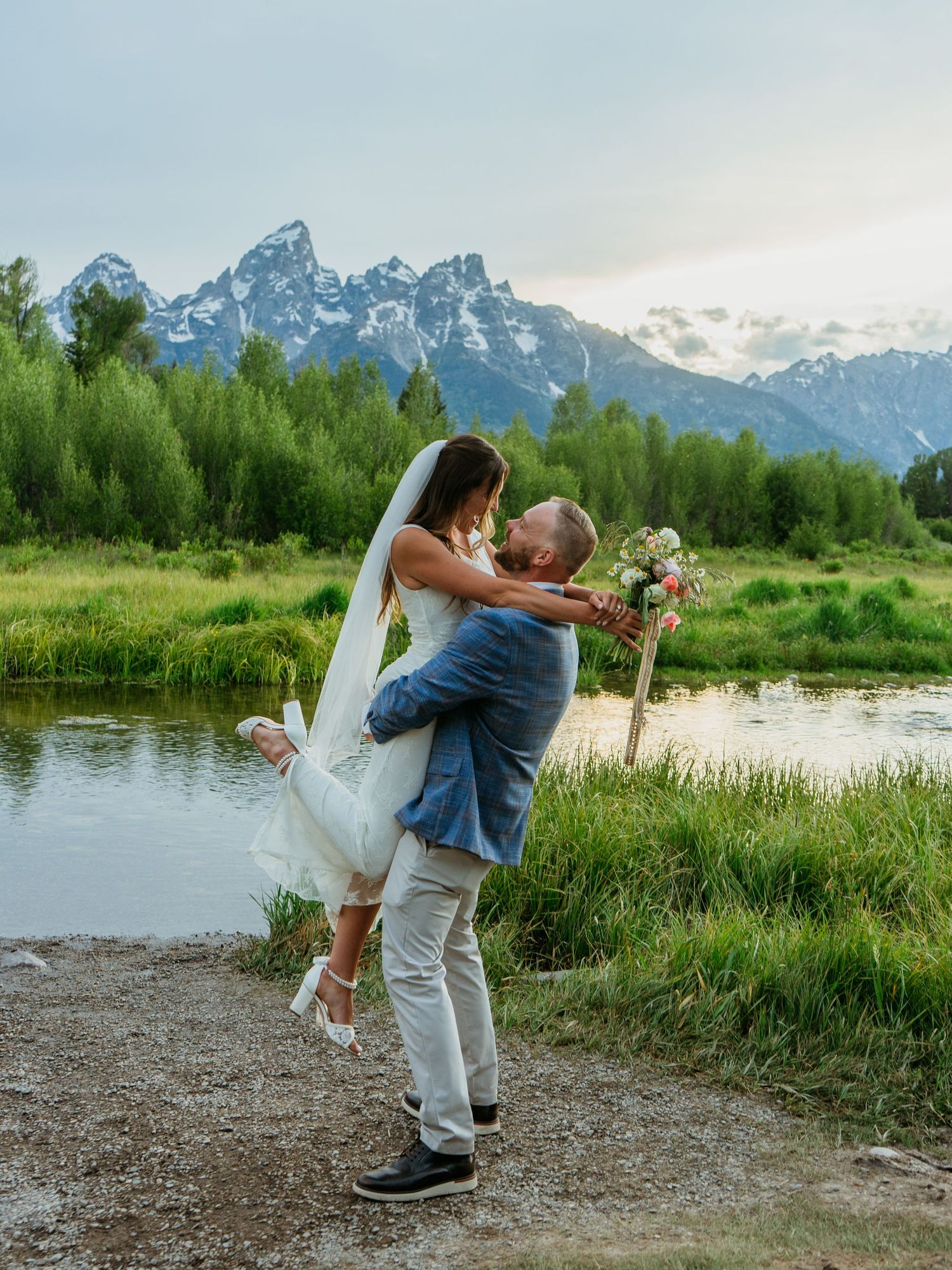 Groom lifts bride in wedding attire by a serene mountain lake with the Grand Tetons and lush greenery in the background.
