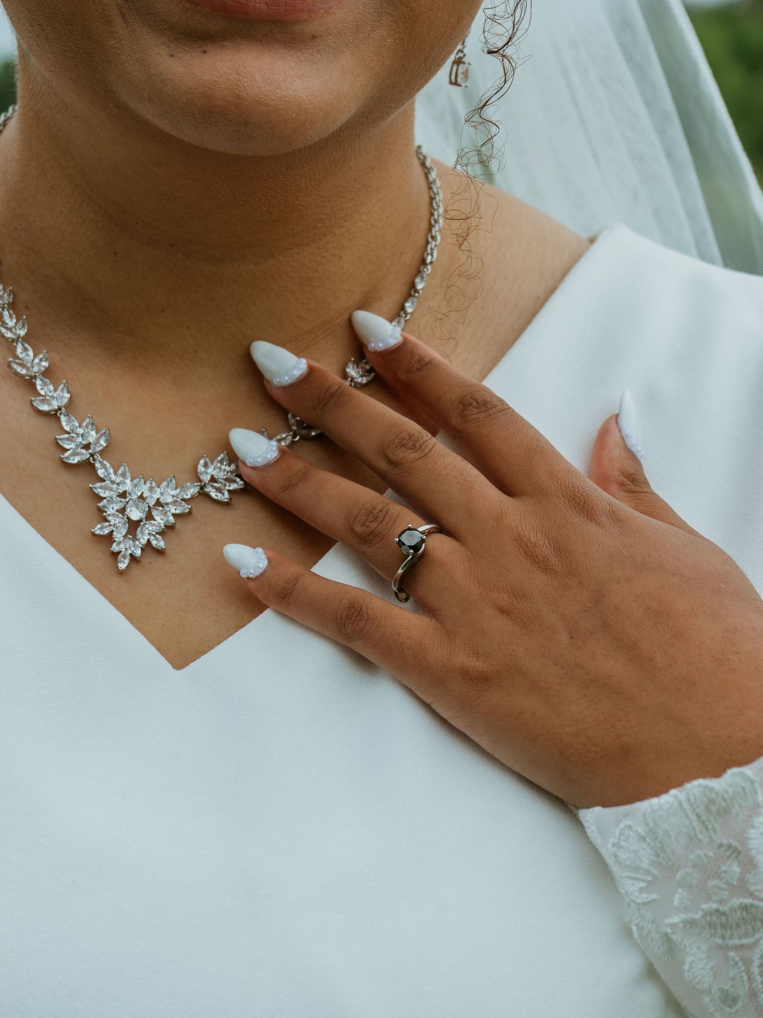 Bride wearing a crystal statement necklace, diamond ring, and white manicure against a white wedding dress.