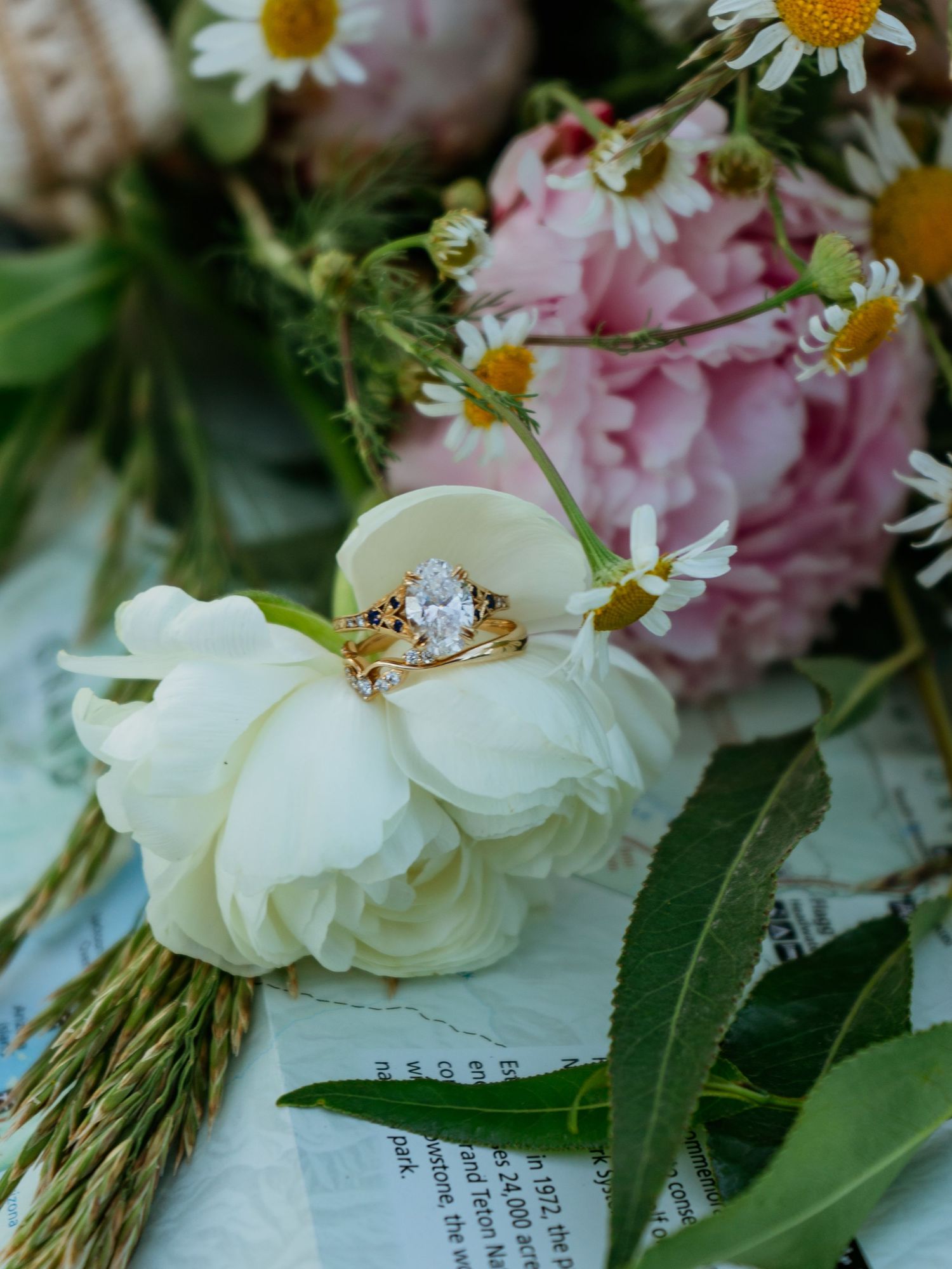 Gold engagement and wedding rings resting on a white ranunculus surrounded by pink peonies and daisies with a Teton National Park map.