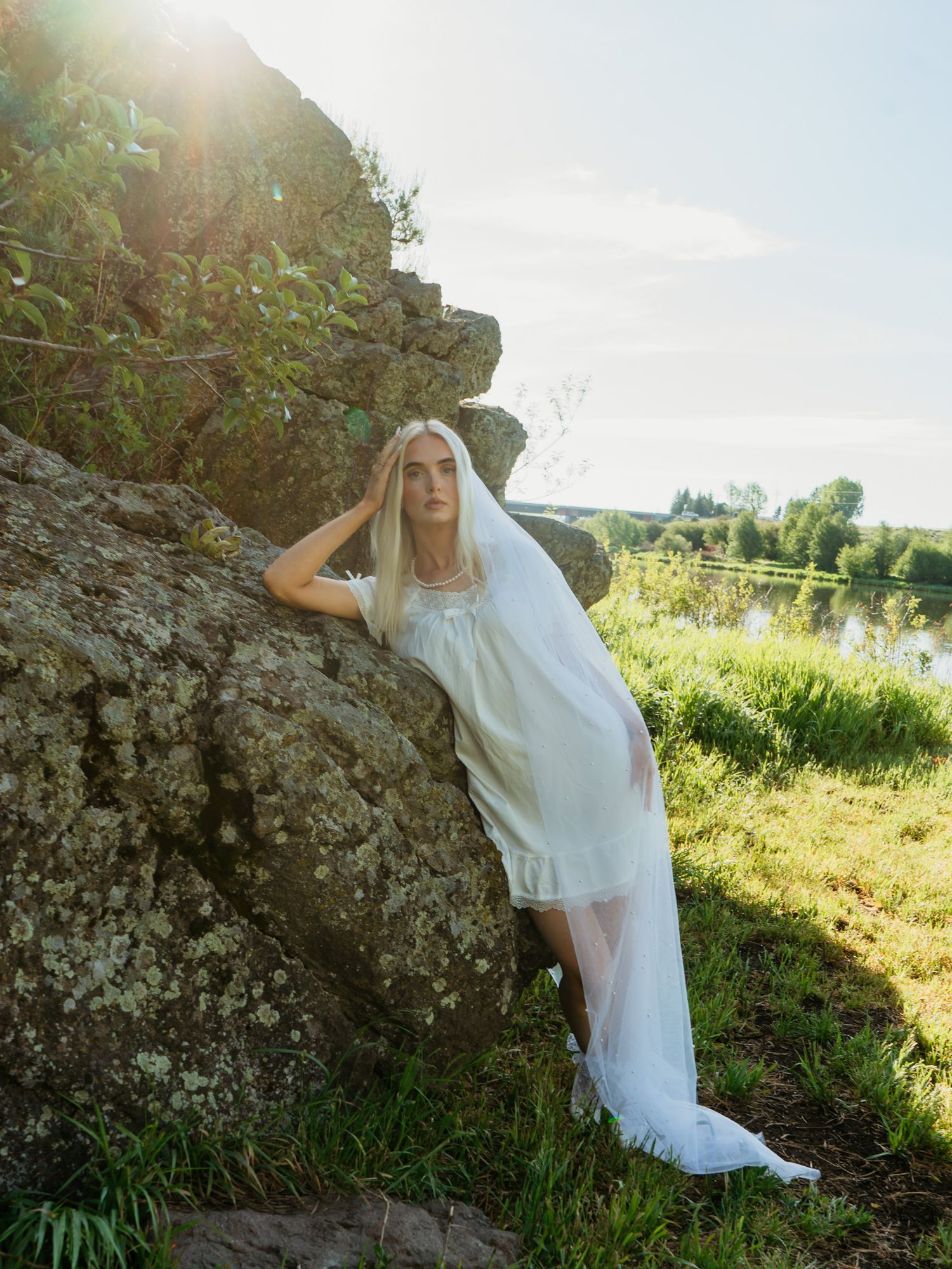 A blonde woman in an elopement dress and veil leans against a mossy rock outdoors near a river in sunlight.