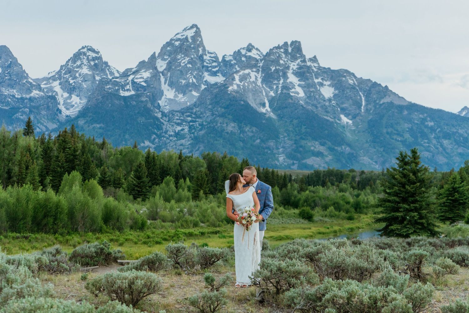 Couple embraces in wedding attire with Grand Teton mountains and lush green forest backdrop in Wyoming.