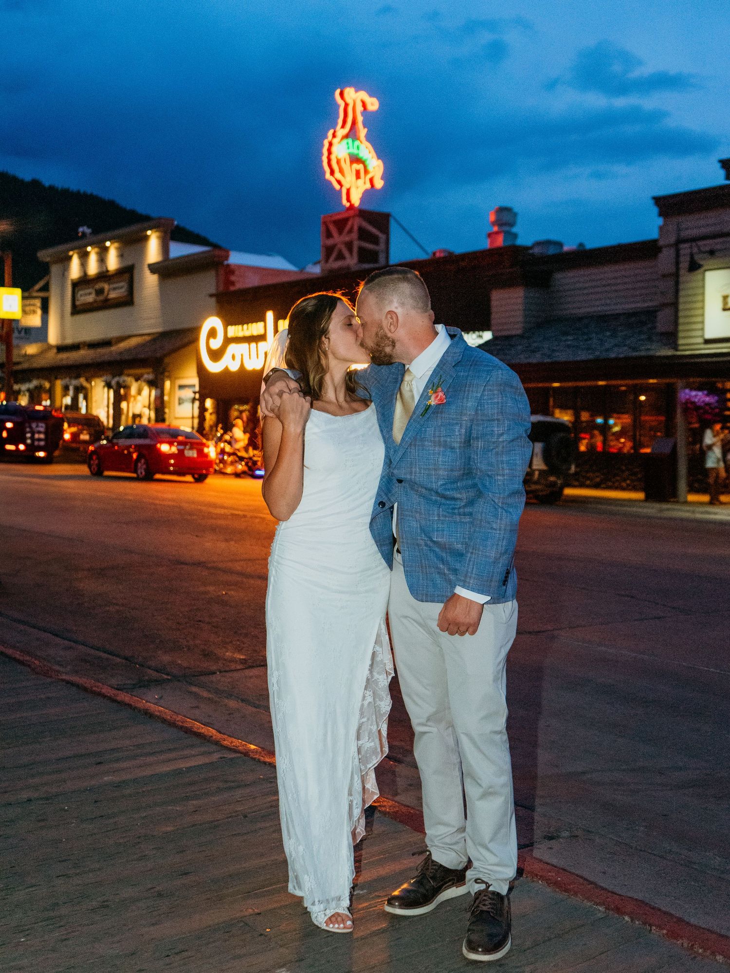 Bride and groom sharing a kiss on a street at dusk with a neon cowboy sign glowing behind them.