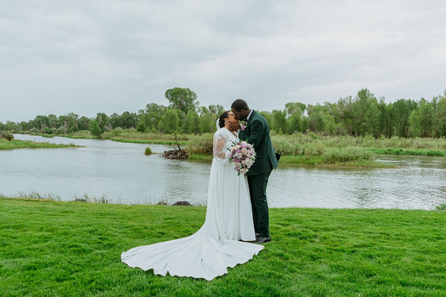 Bride and groom share a kiss by a scenic river on their wedding day, surrounded by lush green grass and trees.