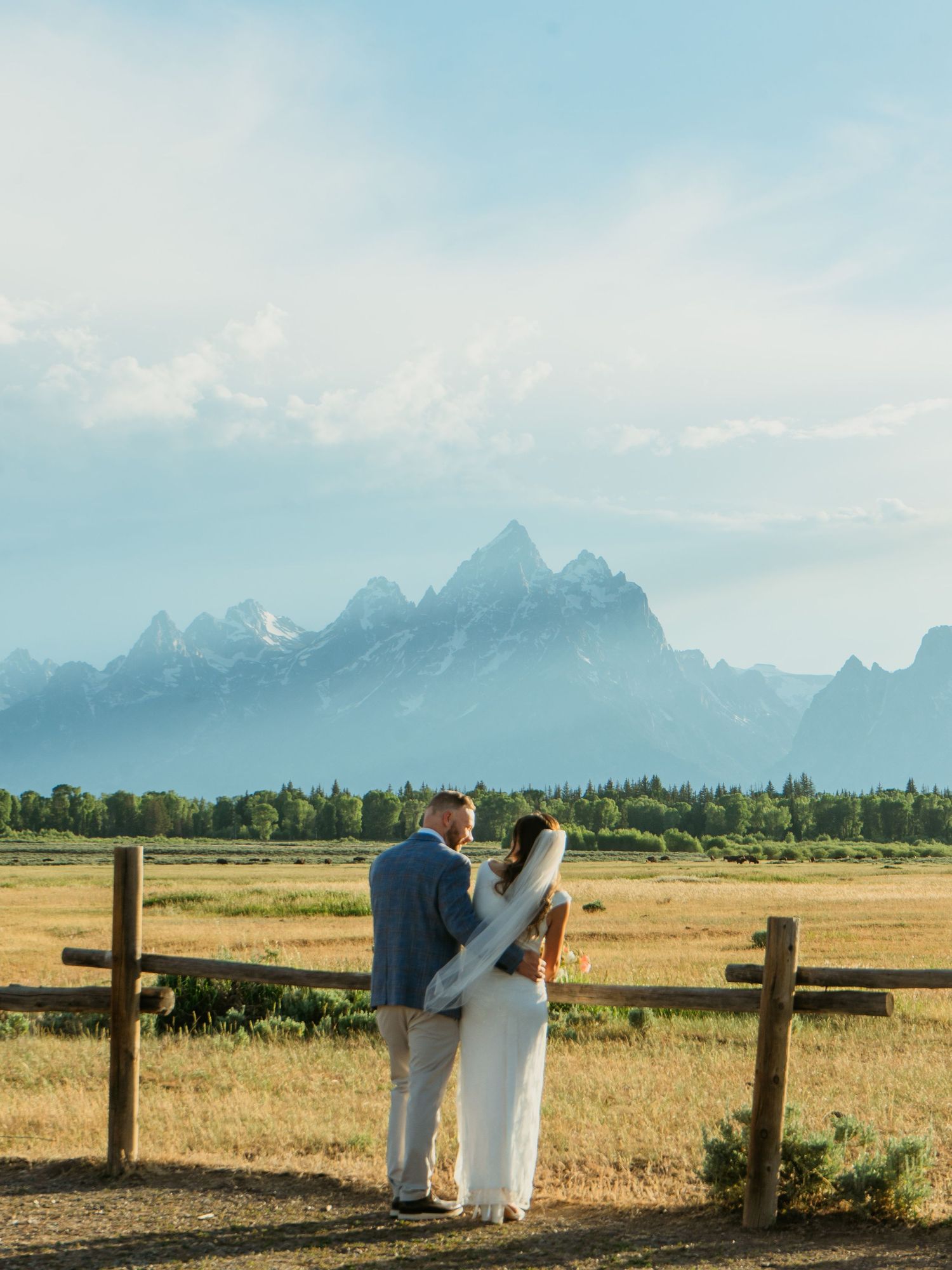 Couple elopes at Grand Teton National Park, standing by a rustic fence with majestic snow-capped mountains in the background.