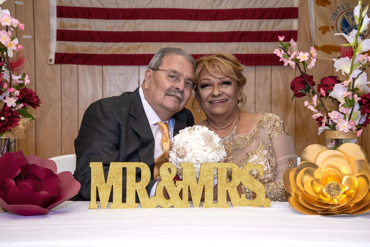 Smiling couple poses behind a glittery MR & MRS sign at their wedding reception, decorated with floral arrangements.