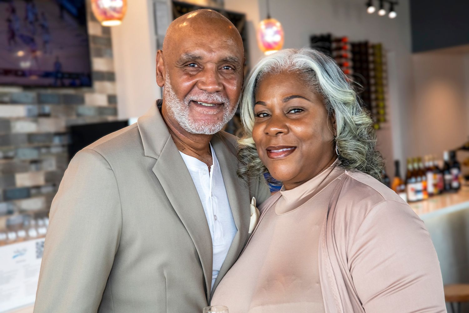 A smiling couple poses together in a restaurant, the man in a gray suit and woman in a pink top with gray hair.