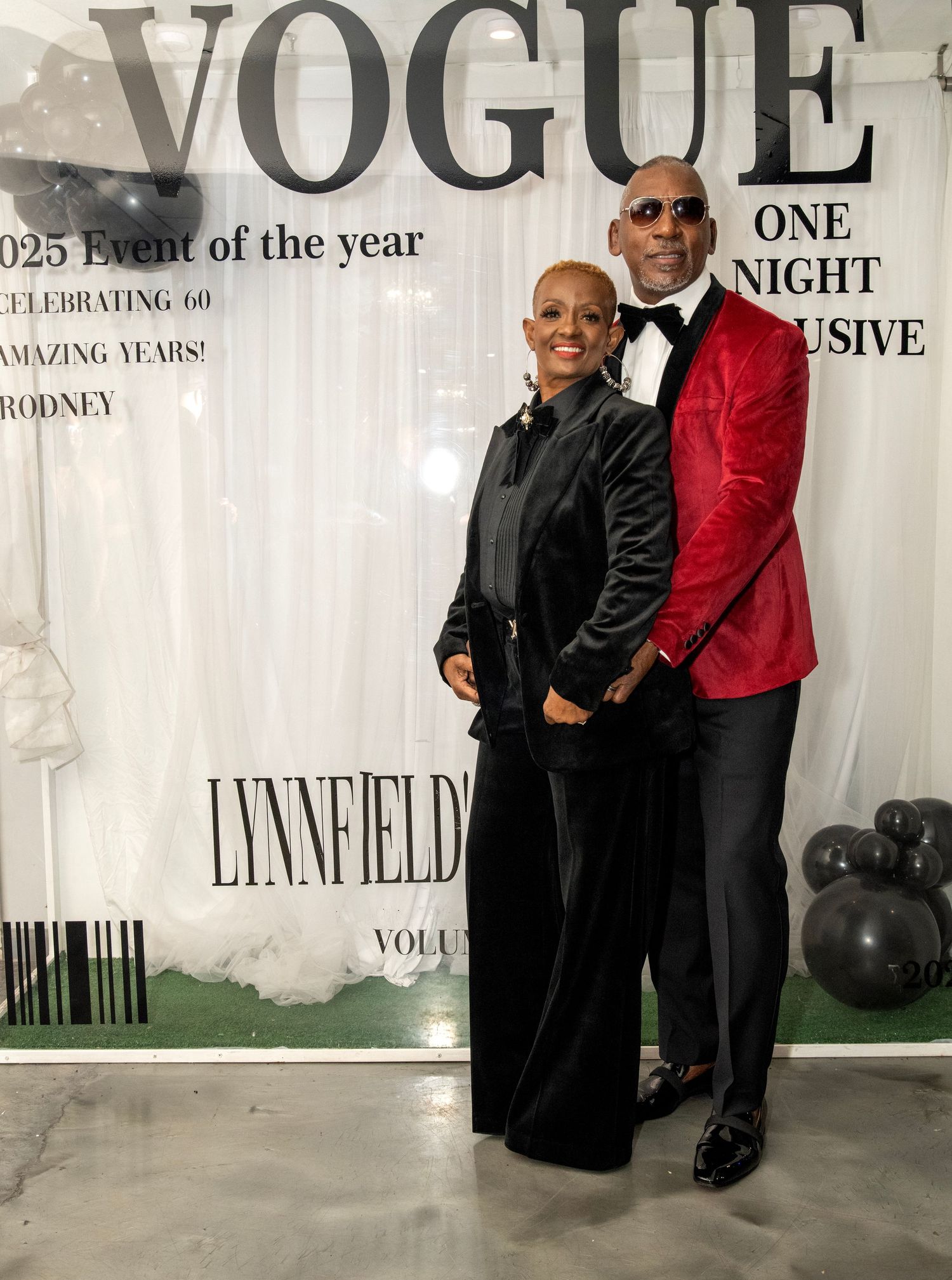 Couple posing at a Vogue-themed 60th birthday celebration event, dressed in elegant black and red formal attire.