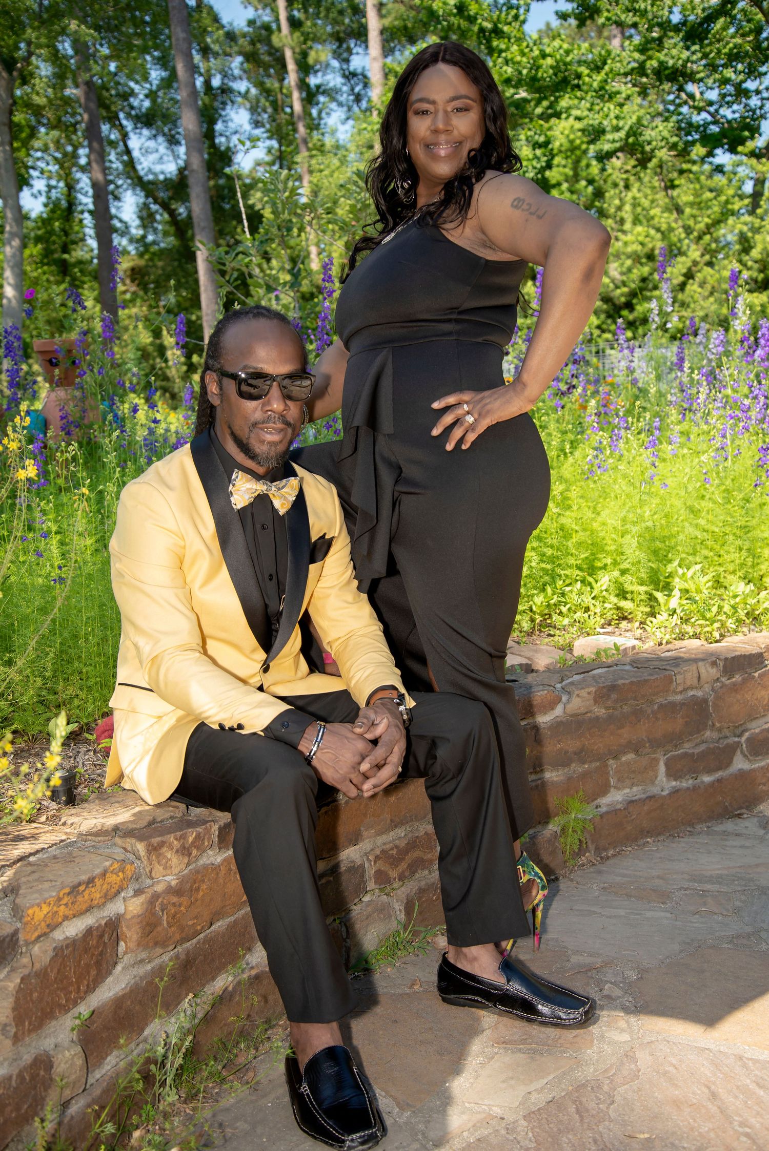 Elegant couple posing outdoors, man in yellow blazer and woman in black jumpsuit, surrounded by lush garden greenery.