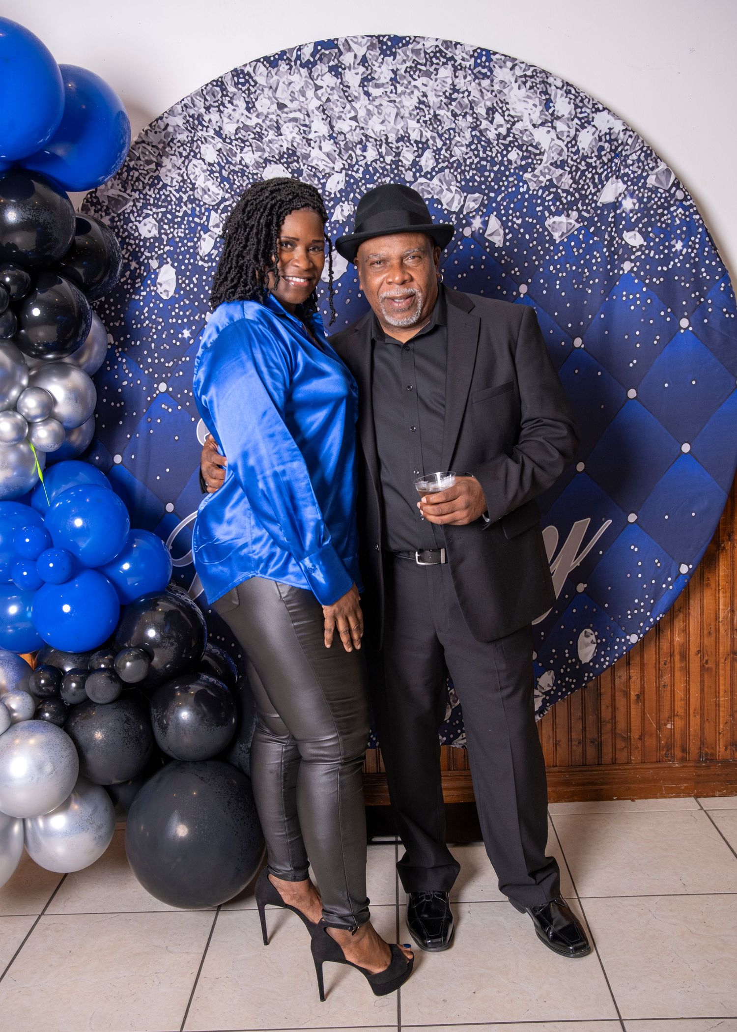 Two people posing at a blue and black balloon backdrop at a formal celebration event.