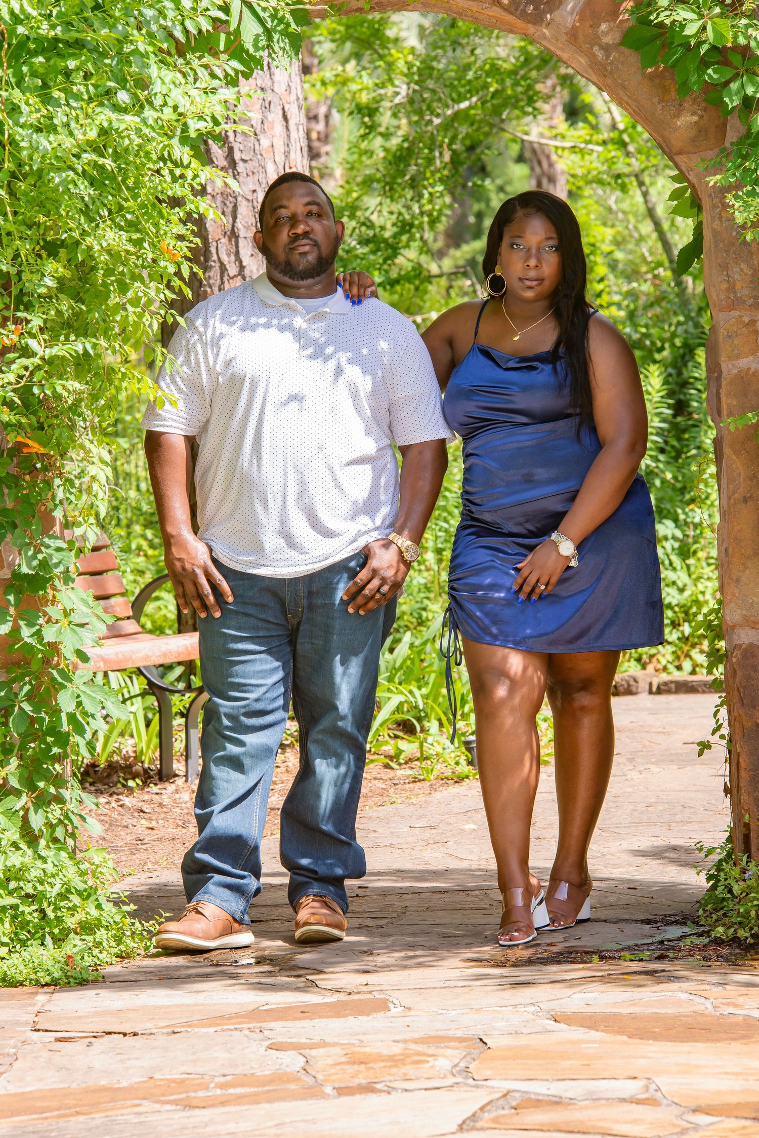 A couple walks hand-in-hand through a lush garden archway, man in white shirt and jeans, woman in blue dress.