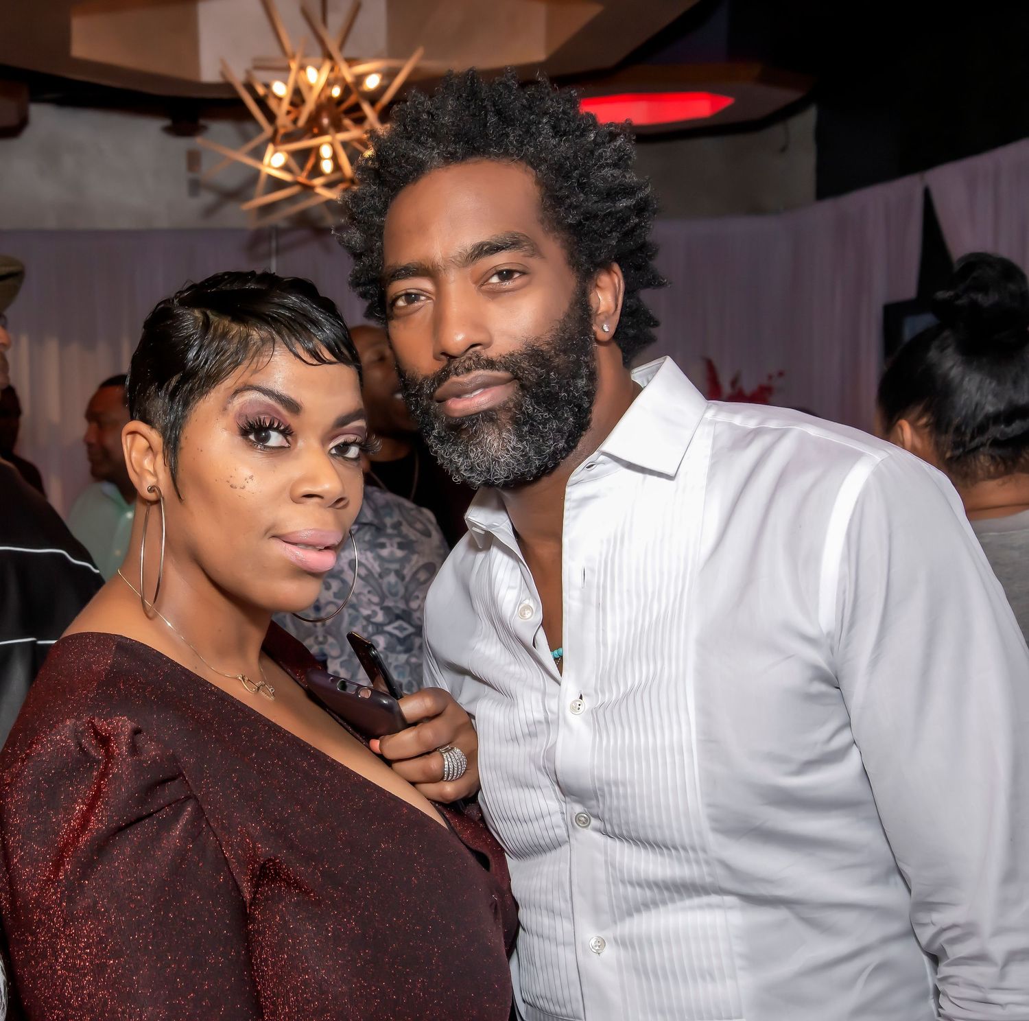 A couple poses together at an indoor event, woman in sparkly top and man in white dress shirt, warm ambient lighting.
