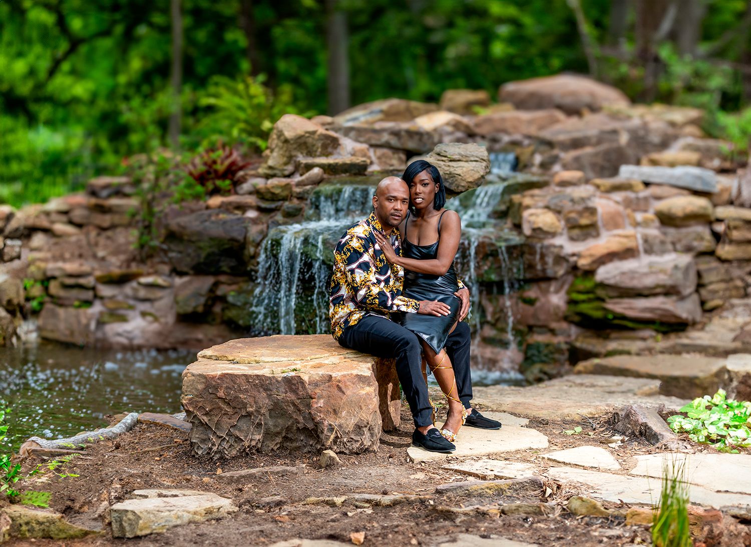 Couple sitting on rocks in front of a stone waterfall surrounded by lush green trees in an outdoor setting.