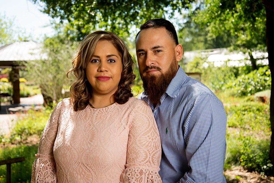 A couple poses outdoors in a lush garden, woman in pink lace top and man in blue striped shirt.