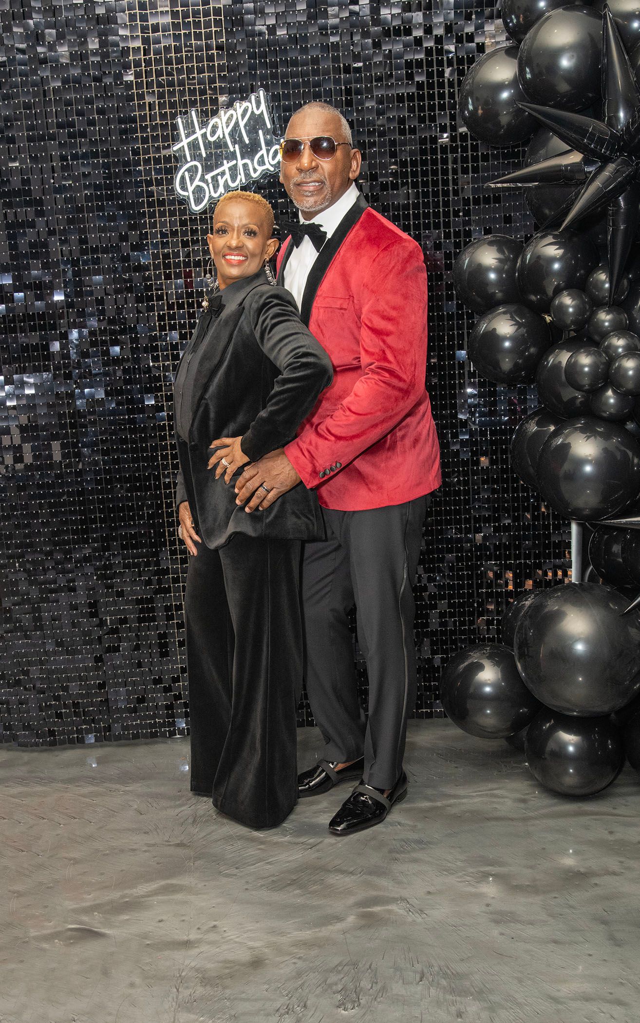 Elegant couple posing at a Happy Birthday celebration with black balloons, woman in black gown and man in red blazer.