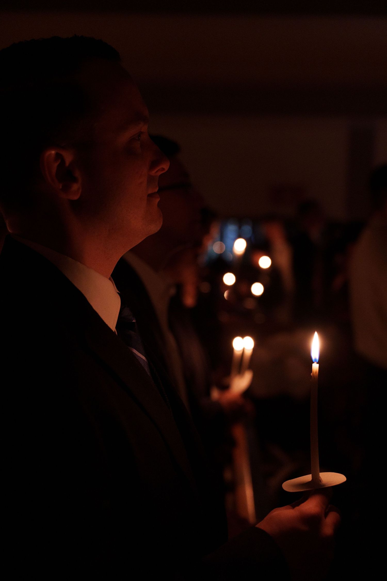 Man in dark suit holds lit candle during solemn candlelight ceremony or religious service in dimly lit venue.